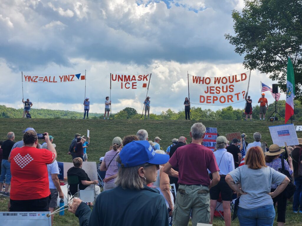 Several signs against a cloudy sky with protesters in the foreground. The signs say Yinz - All Yinz, Unmask ICE, and Who Would Jesus Deport.