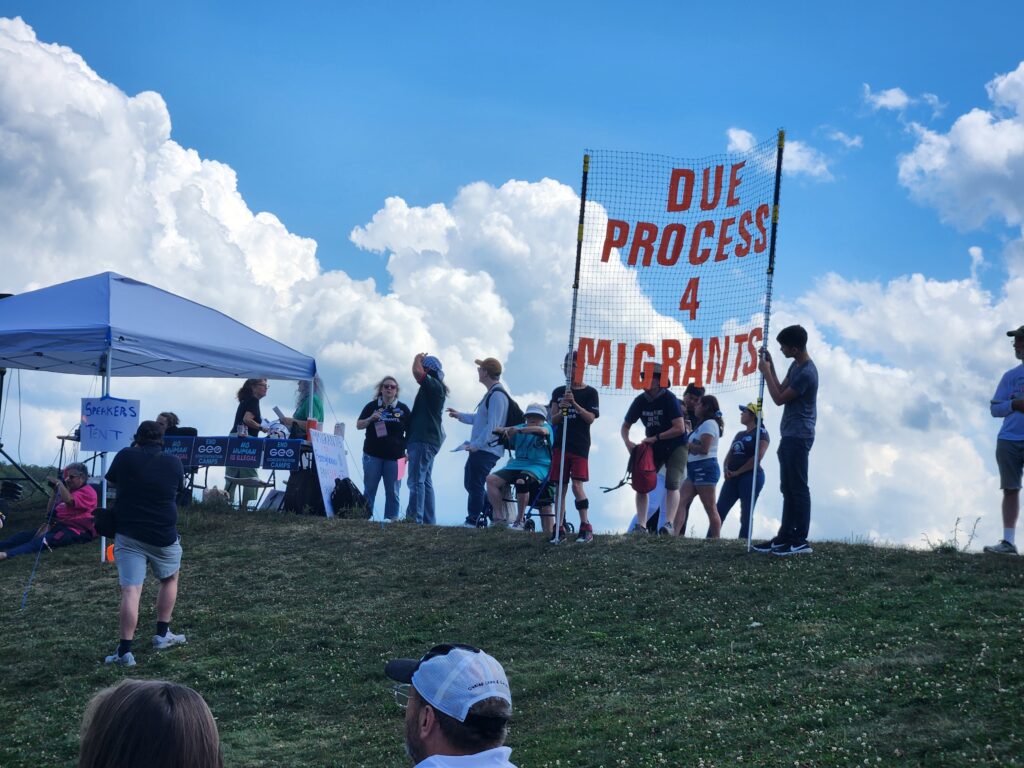 Several people on a hill holding a sign with red letters. The sign reads Due Process 4 Migrants.